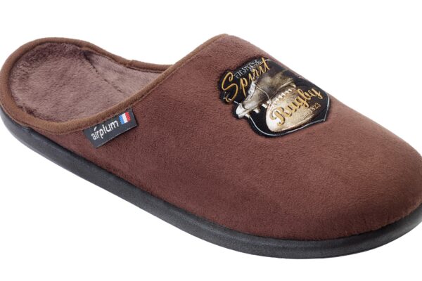 chausson de type mule en couleur marron. Il est conçu avec une doublure douce et confortable à l'intérieur. Sur le dessus, il y a un écusson décoratif noir et doré comportant l'inscription "Spirit Rugby" et un motif lié au rugby, comme une balle ou un casque. Sur le côté, une étiquette affiche le logo de la marque "airplum" accompagné d'un petit drapeau français, indiquant probablement que ce produit est fabriqué en France ou associe la marque à une identité française. La semelle semble être noire, robuste et adaptée pour un usage intérieur.